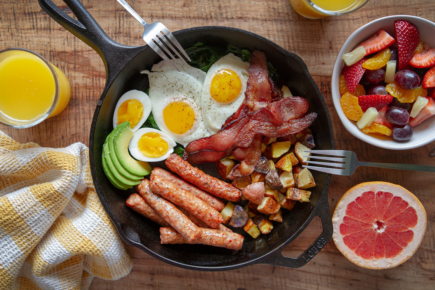 A cast iron skillet is filled with over easy eggs, skillet potatoes, sausage links, and bacon. There is a bowl of fruit and a glass of orange juice beside it.