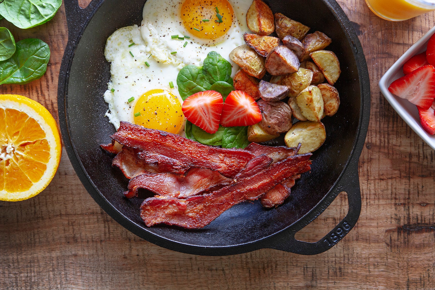 Several slices of cooked Pederson's Farms Applewood Smoked Bacon are shown in a cast iron skillet. Beside the bacon are golden breakfast potatoes, two eggs, fresh spinach, and halved strawberries.