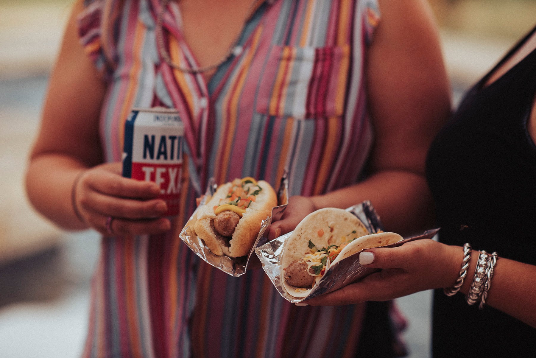 Closeup image of two girls standing side by side holding a beer brat in a bun and in a tortilla.