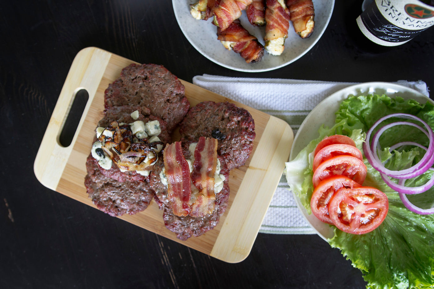 Overhead view of cooked hamburger patties on a wooden cutting board with organic bacon strips laid over the top.