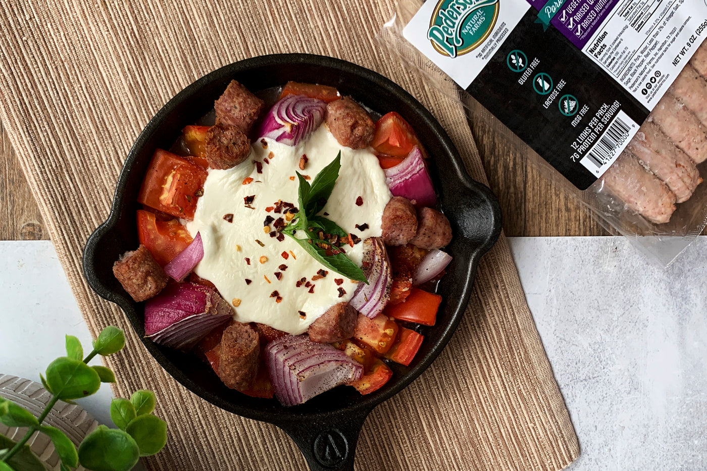 Image of a Burrata Breakfast Skillet - Pederson's Fully Cooked Mild Breakfast Sausage Links with largely chopped tomato & onion, topped with one burrata sprinkled with basil & red pepper flakes. The dish is in a cast iron skillet with wood background.