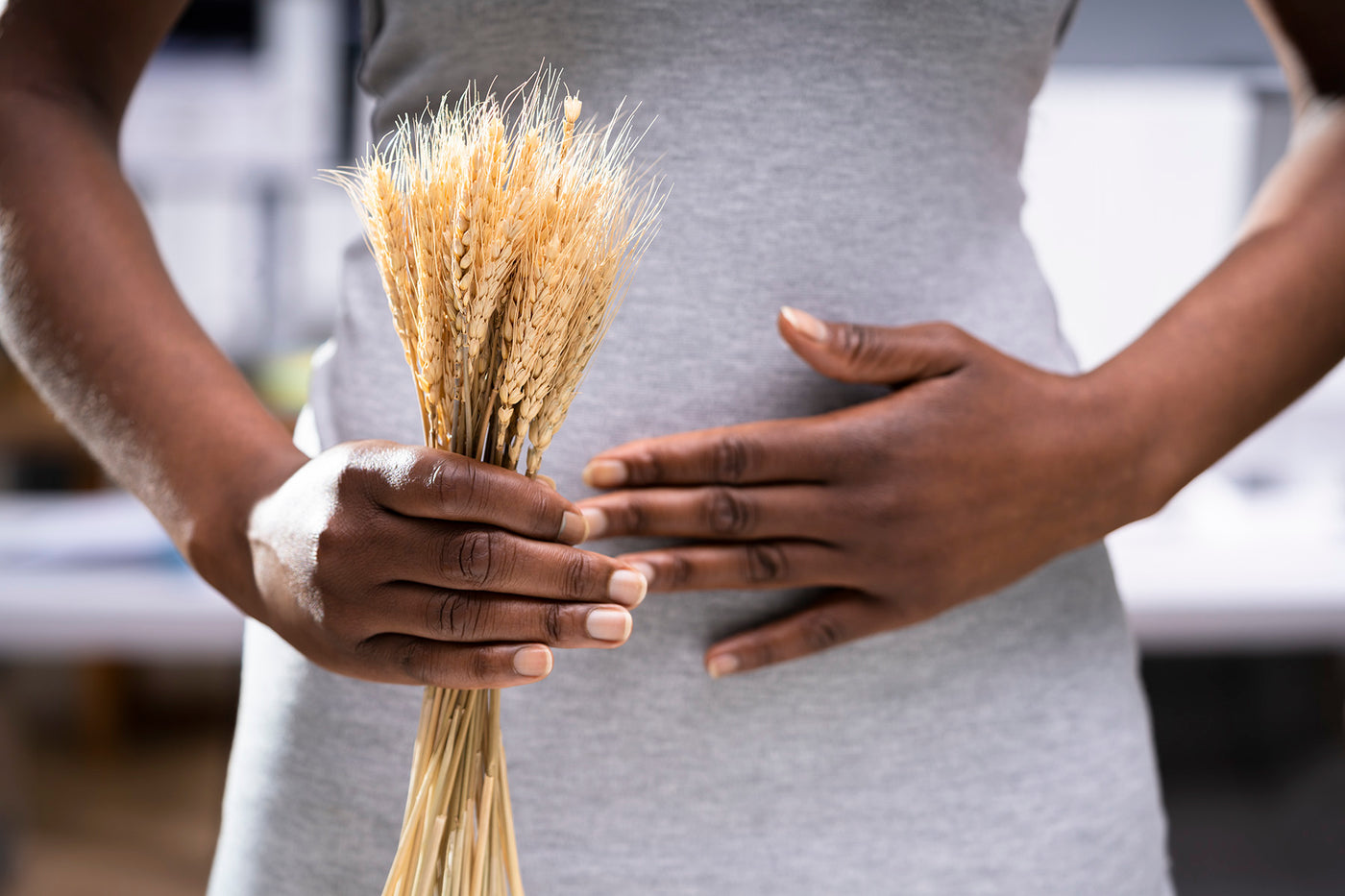 The midsection of a woman is shown. In one hand, she is holding a bunch of wheat. The other hand is over her stomach indicating that she has celiac disease.