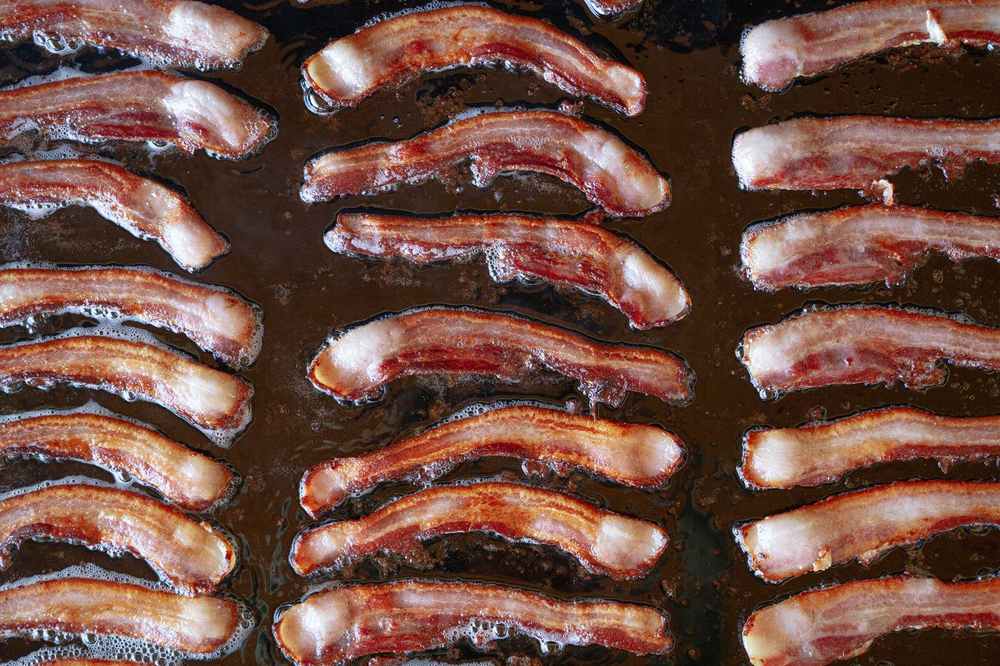 Several slices of uncured bacon are frying on a black cook top.