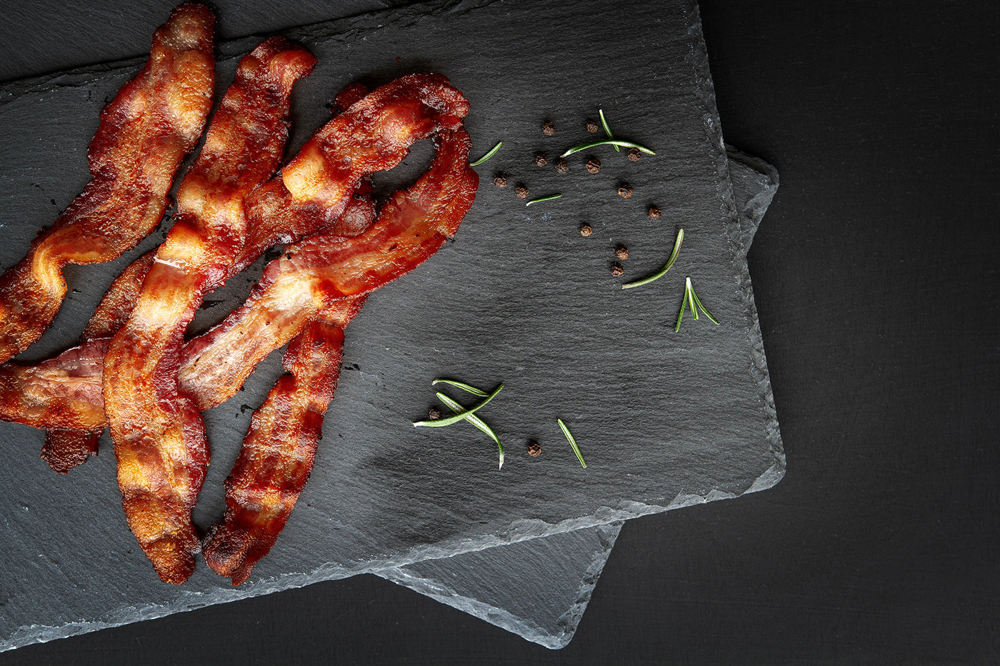4 slices of cooked bacon sit on a black slate with loose herbs around the edges.