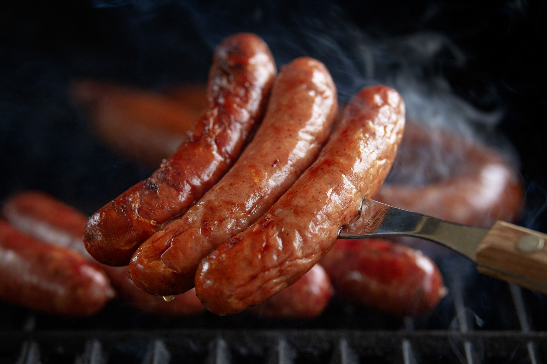 3 Uncured Sausage Links are shown on a large fork over the grates of a grill as smoke rises in the background.