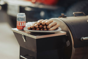 A plate of uncured beer bratwurst sits on the edge of a BBQ grill with a Native Texan Pilsner can of beer in the background.