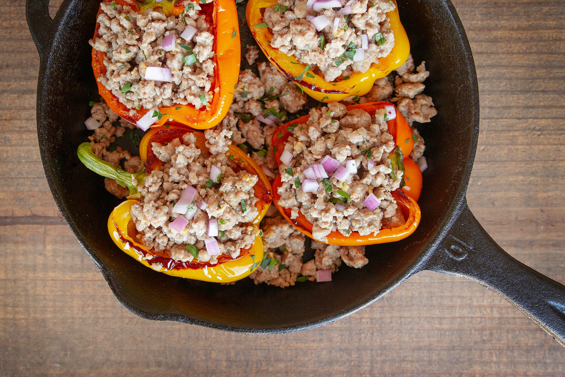 Four yellow and orange bell peppers are stuffed with cooked ground pork and onions. They sit inside a cast iron skillet on a wooden tabletop.