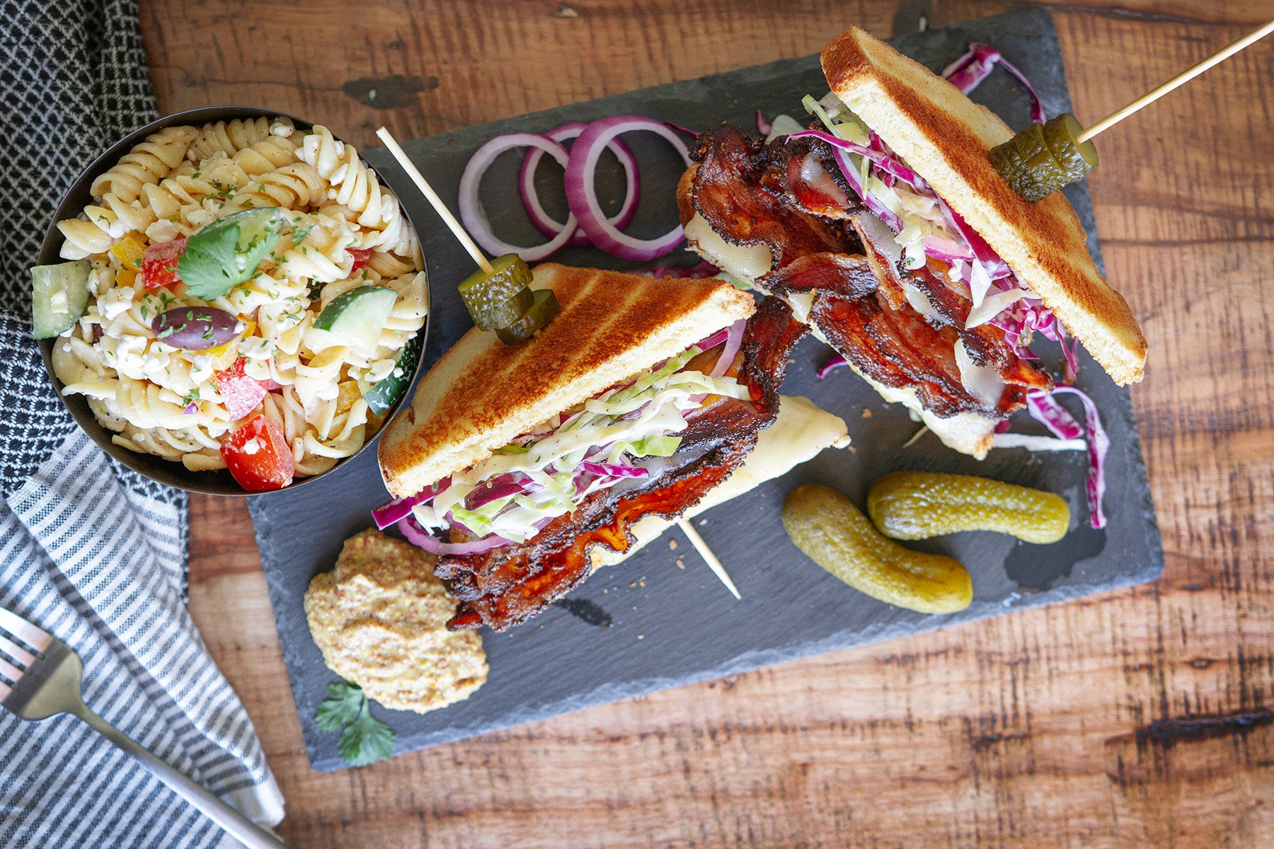 A pastrami bacon sandwich on toasted bread topped with a cabbage slaw and mustard. A small bowl of pasta salad sits near the sandwich, as well as small dill pickles.