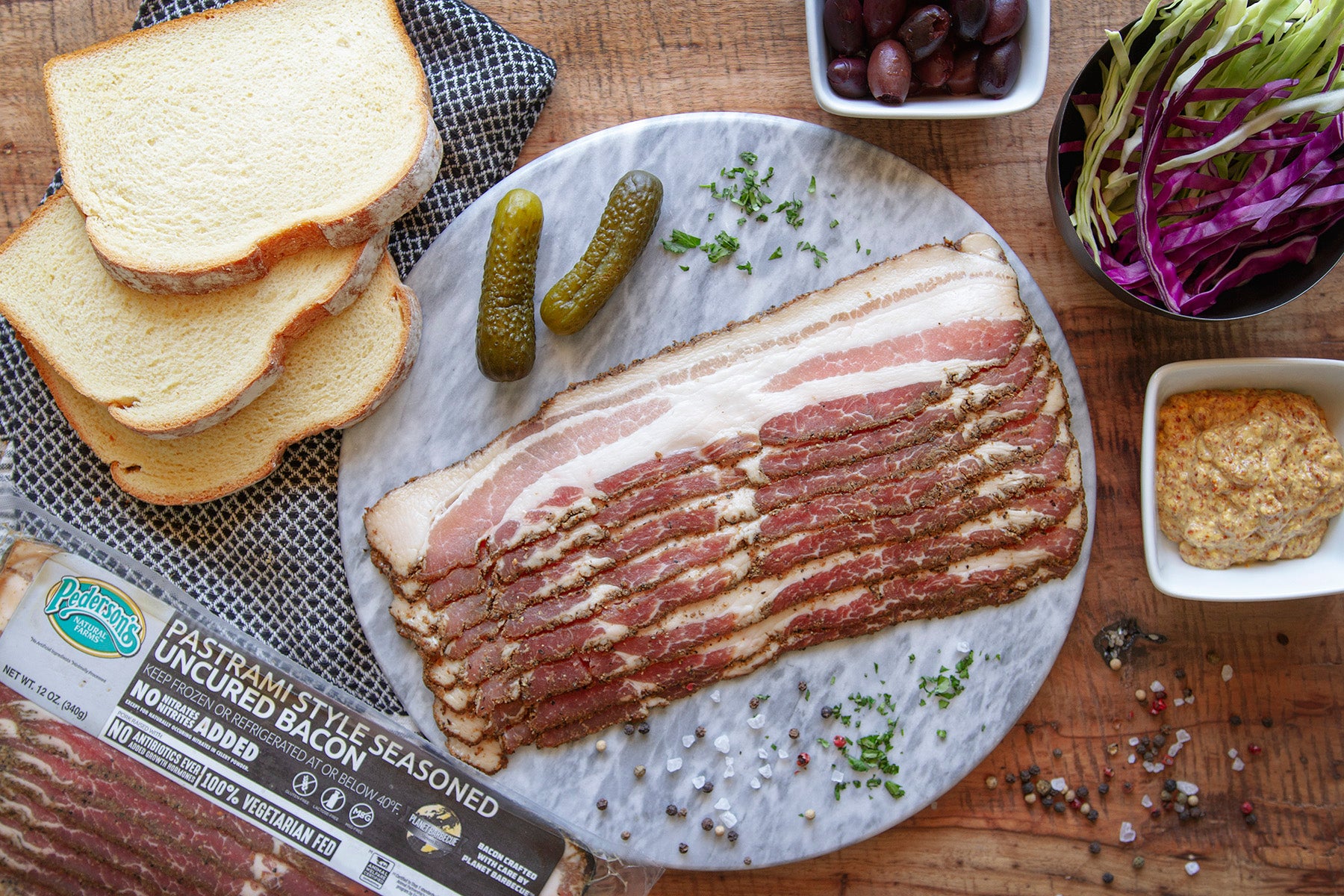 Raw pastrami bacon sits on a marble slab surrounded by fresh bread, pickles, cabbage, and mustard