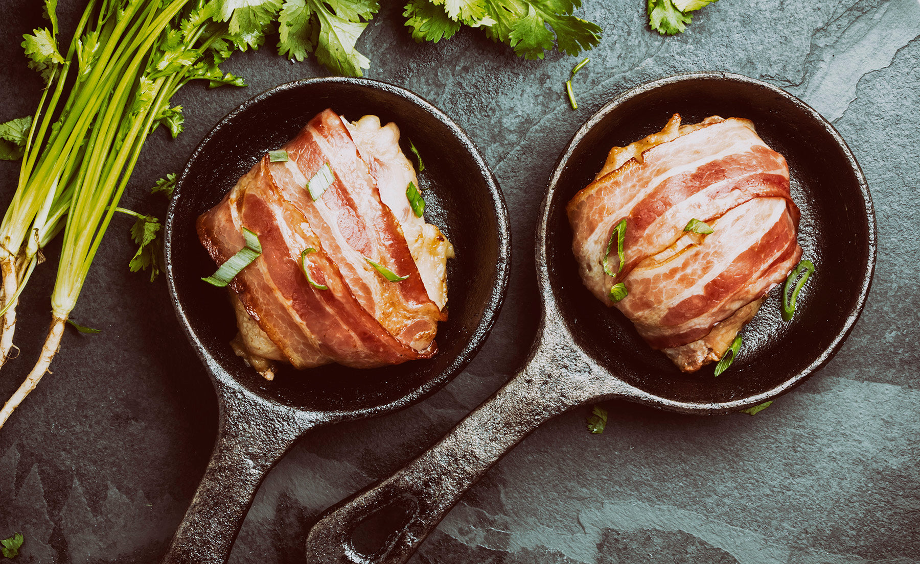 Two bacon wrapped chicken breasts sit in two miniature skillets with chopped herbs surrounding it. 
