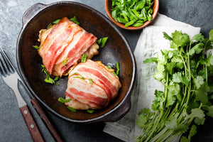 Two bacon wrapped chicken breasts sit in a cast iron skillet with diced herbs around it. 