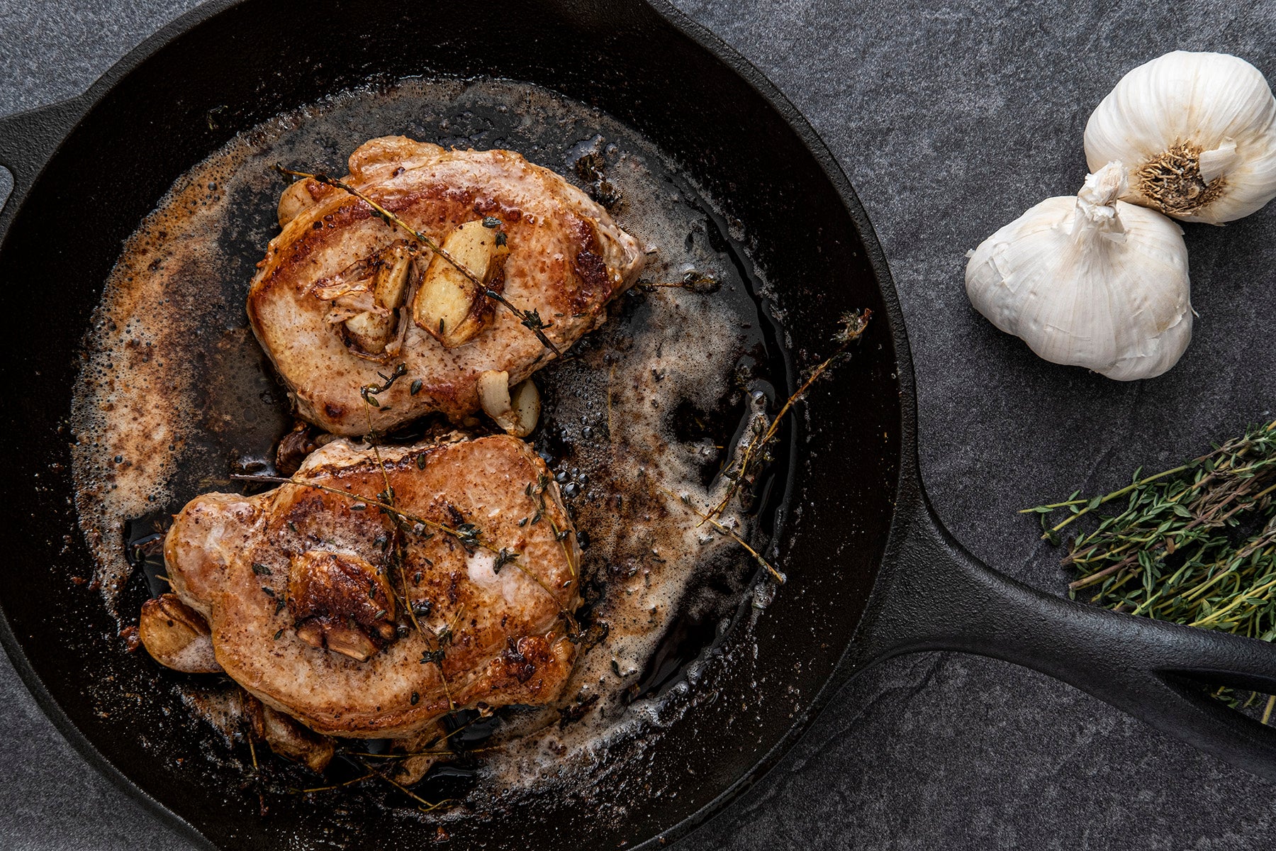 Two perfectly browned thick cut pork chops are shown in a cast iron skillet with roasted garlic cloves and herbs on top.