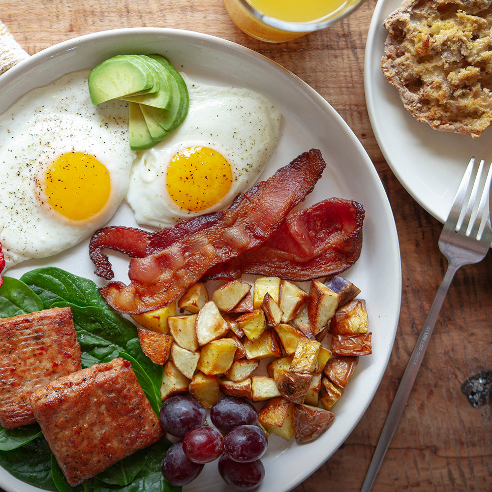 Apple wood smoked bacon breakfast plate lifestyle image featuring two sunny side up eggs, avocado slices, Pederson's Farms sausage patties, home fries, and purple grapes. the plate is positioned on a wooden table with a fork, a glass of OJ and another plate of food.