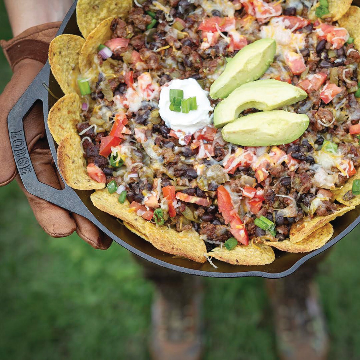 A person holding a skillet filled with a Organic Ground Beef taco salad topped with three Avocado slices and a scoop of sour cream surrounded by a ring of circle chips.