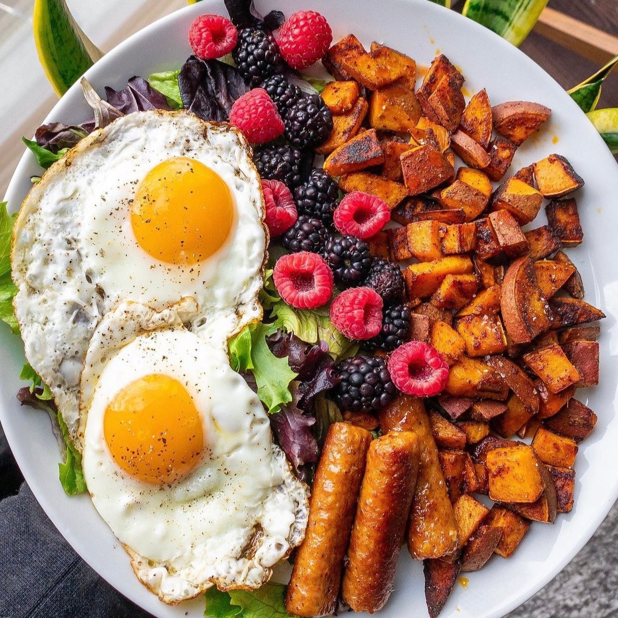 Protein packed lunch served with diced sauteed sweet potatos, small salad topped with two sunny-side-up eggs, mix of raspberries and blackberries, finished with three fully cooked mild breakfast sausage links.